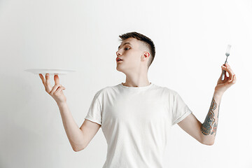 Image showing Young smiling attractive guy holding empty dish and fork isolated on grey background.