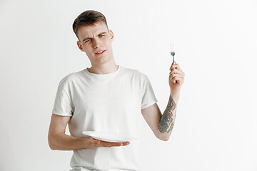 Image showing Young sad attractive guy holding empty dish and fork isolated on grey background.