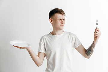 Image showing Young sad attractive guy holding empty dish and fork isolated on grey background.