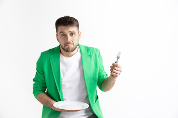 Image showing Young sad attractive guy holding empty dish and fork isolated on grey background.