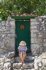 Image showing Beautiful young female tourist woman wearing big straw hat using moile phone, sitting in front of white vinatage wooden door and textured stone wall at old Mediterranean town