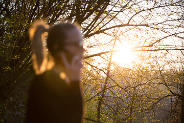 Image showing Backlit defocused rear view of young woman talking on cell phone outdoors in park at sunset. Girl holding mobile phone, using digital device, looking at setting sun