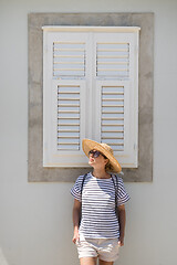 Image showing Beautiful young female tourist woman wearing sun hat, standing and relaxing in front of vinatage wooden window in old Mediterranean town while sightseeing on hot summer day