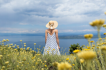 Image showing Rear view of young woman wearing striped summer dress and straw hat standing in super bloom of wildflowers, relaxing while enjoing beautiful view of Adriatic sea nature, Croatia