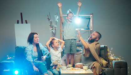 Image showing Happy family watching projector, TV, movies with popcorn in the evening at home. Mother, father and kids spending time together.