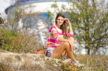 Image showing A girl sits on a hillock, in the background there is a beautiful structure and bushes, the girl looks into the frame