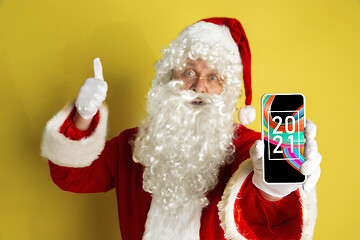 Image showing Stylish Santa Claus in traditional costume with modern device pointing on 2021 on yellow studio background