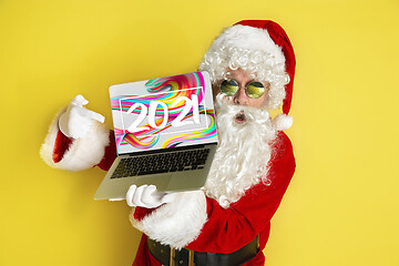 Image showing Stylish Santa Claus in traditional costume with modern device pointing on 2021 on yellow studio background