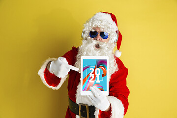 Image showing Stylish Santa Claus in traditional costume with modern device pointing on 2021 on yellow studio background