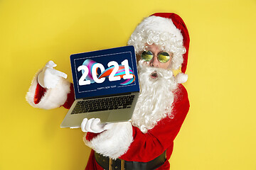 Image showing Stylish Santa Claus in traditional costume with modern device pointing on 2021 on yellow studio background