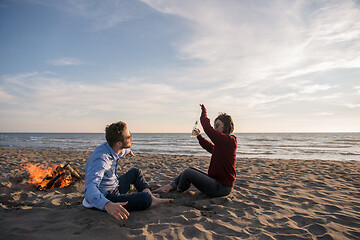 Image showing Young Couple Sitting On The Beach beside Campfire drinking beer