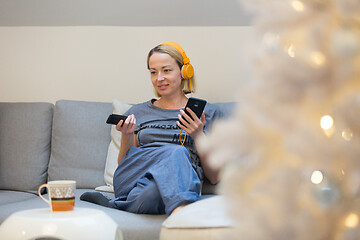 Image showing Young cheerful woman sitting indoors at home living room sofa watching TV, using social media on phone for video chatting and staying connected with her loved ones