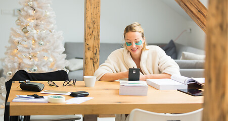 Image showing Young businesswoman wearing cosy warm bathrobe and cosmetic eye patches working remotly from home in winter Christmas time during 2020 corona virus pandemic. Work from home, selter in place, concept