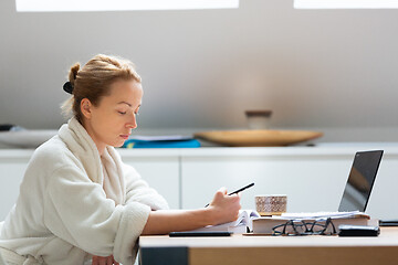 Image showing Young female entrepreneur wearing cosy warm bathrobe working remotly from home in winter Christmas time during 2020 corona virus pandemic. Work from home, selter in place, concept.