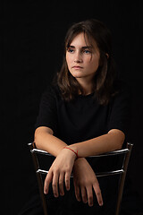 Image showing Portrait of a young girl sitting on a high chair and folded her hands on the back of a chair, black background