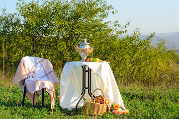 Image showing On the field under the bushes is a table with a samovar and fruit, next to it is a chair