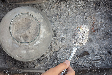 Image showing Storage of wood ash in a glass jar, next to a hand holds a spoon with fresh ash