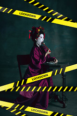 Image showing Young japanese woman as geisha isolated on dark green background. Drinking cola and eating popcorn behind limiting message lockdown on the screen