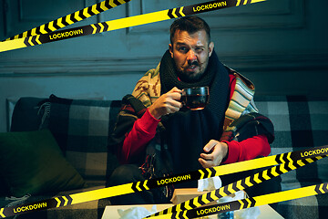 Image showing Young man wrapped in a plaid looks sick, ill, sneezing and coughing sitting on sofa at home behind limiting tapes with word Lockdown