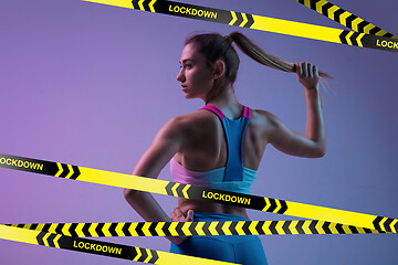 Image showing Young sportive woman posing on gradient neon background behind limiting tapes with word Lockdown