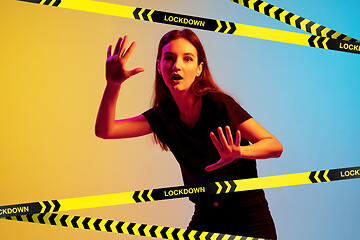 Image showing Young sportive woman posing on gradient neon background behind limiting tapes with word Lockdown