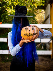 Image showing A girl with her hair covered face in a witch costume is holding a pumpkin with a painted face and a witch\'s cap