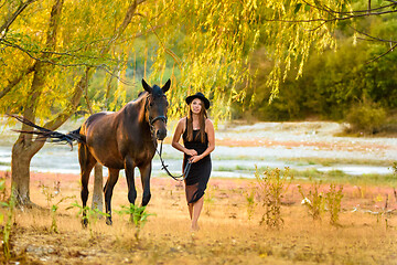 Image showing A girl in a beautiful black dress and a black hat walks with a horse across the field