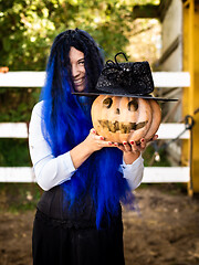 Image showing A girl dressed as a witch with blue hair is holding a pumpkin with a painted face and a witch\'s cap