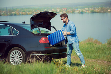 Image showing Handsome smiling man going to vacations, loading his suitcase in car trunk