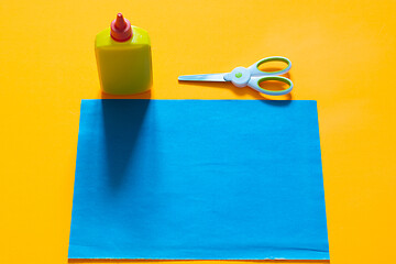 Image showing On a yellow background lies a sheet of blue colored paper, next to it there is a tube of glue and scissors