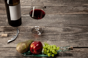 Image showing A bottle and a glass of red wine with fruits over dark stone background