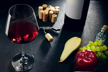 Image showing A bottle and a glass of red wine with fruits over dark stone background