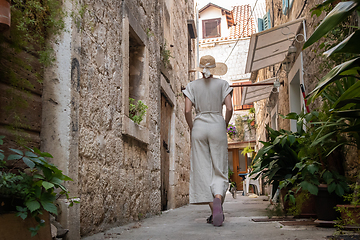 Image showing Rear view of beautiful blonde young female traveler wearing straw sun hat sightseeing and enjoying summer vacation in an old traditional costal town at Adriatic cost, Croatia