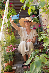 Image showing Beautiful female tourist wearing big straw sun hat and shorts sitting and relaxing on old stone house stairs during summer travel on Mediterranean cost on hot summer day and taking selfie