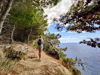 Image showing Young active feamle tourist wearing small backpack walking on coastal path among pine trees looking for remote cove to swim alone in peace on seaside in Croatia. Travel and adventure concept