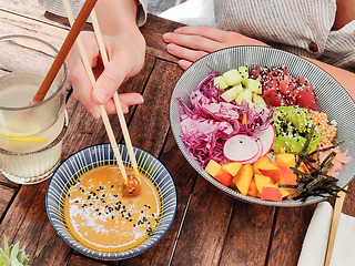 Image showing Woman eating tasty colorful healthy natural organic vegetarian Hawaiian poke bowl using asian chopsticks on rustic wooden table. Healthy natural organic eating concept