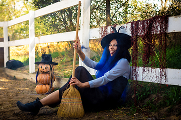 Image showing A girl dressed as a witch sits by a fence with a broom in her hands and shows her tongue