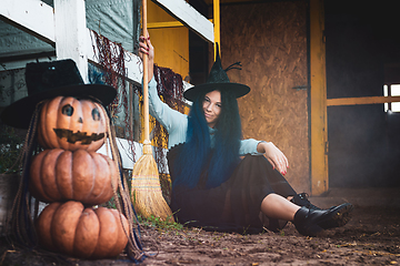 Image showing A girl dressed as a witch sits by a fence with a broom in her hands, in the foreground there is a scary figure of pumpkins