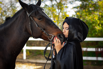 Image showing Girl in a black cloak hugs and kisses a horse