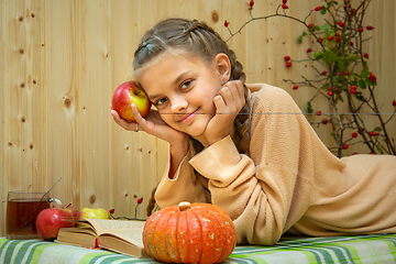 Image showing A girl lying down reads a book, a pumpkin and apples lie nearby, a girl joyfully looks into the frame