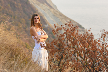 Image showing A beautiful girl stands on a rocky seashore and hugs wildflowers