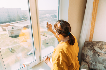 Image showing Woman Of Fifty In Yellow Sweater And Jeans Washes Dusty Window In Apartment. 50 Year Old Woman Cleans Windows From Stains Using Rag And Spray Cleaner. Caucasian Elderly Woman Is Cleaning House, Doing