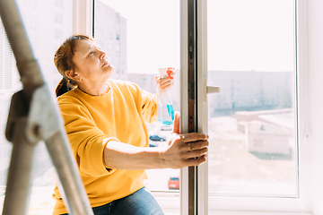 Image showing Woman Of Fifty In Yellow Sweater And Jeans Washes Dusty Window In Apartment. 50 Year Old Woman Cleans Windows From Stains Using Rag And Spray Cleaner. Caucasian Elderly Woman Is Cleaning House, Doing