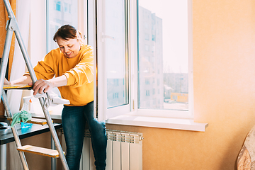 Image showing Smiling Caucasian Woman Of Fifty In Yellow Sweater And Jeans Washes Dusty Window In Apartment. 50 Year Old Woman Cleans Windows From Stains Using Rag And Spray Cleaner. Elderly Woman Is Cleaning Hous