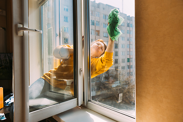 Image showing Caucasian Woman Of Fifty In A Yellow Sweater And Jeans Washes A Dusty Window In Apartment. A 50 Year Old Woman Cleans Windows From Stains Using Rag. Elderly Woman Is Cleaning House, Doing Household C