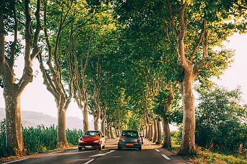 Image showing Cars driving on a country road lined with trees. Bright sunlight at sunset in the evening. Cars on background of French landscape