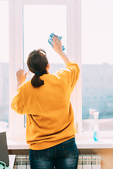 Image showing Woman Of Fifty In Yellow Sweater And Jeans Washes Dusty Window In Apartment. 50 Year Old Woman Cleans Windows From Stains Using Rag And Spray Cleaner. Caucasian Elderly Woman Is Cleaning House, Doing