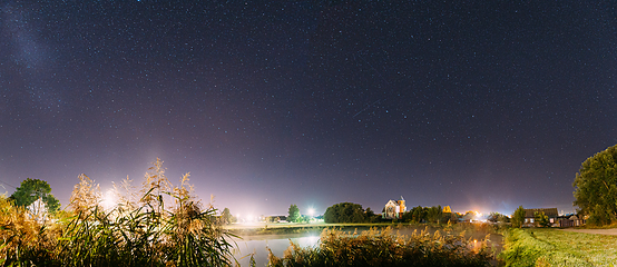 Image showing Panorama Of Evaporation Over River Lake Near Houses In Village. Night Starry Sky Above Lake River With Bright Stars And Meteoric Track Trail. Glowing Stars Above Summer Nature