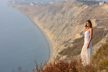 Image showing A beautiful slender girl in a white dress stands on a hill and enjoys a beautiful view of the sunset