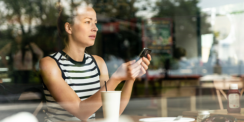 Image showing Thoughtful caucasian woman holding mobile phone while looking through the coffee shop window during coffee break.